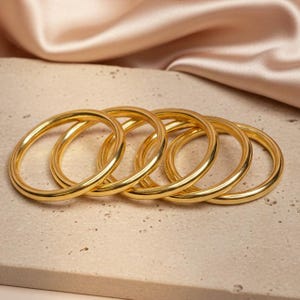 May include: A set of five polished gold-tone metal bangles. Each bangle features a smooth, rounded, circular band design with a reflective finish. The bracelets are arranged in a slightly overlapping row, showcasing their uniform shape and metallic luster.