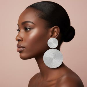 May include: A profile view showcases a person wearing large, silver, disc-shaped earrings. The earrings are composed of two connected circles, with a textured, concentric design. The background is a soft, muted pink.
