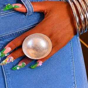May include: A large, circular, silver-toned statement ring with a smooth, reflective, dome-shaped surface. The ring is worn on a finger, accompanied by a stack of thick, polished silver-toned metal bangles on the wrist and blue denim fabric.