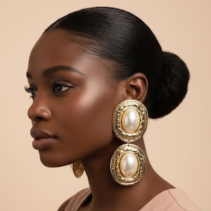 May include: Gold-toned, double-drop earrings with a large, oval, faux pearl center. The earrings have a textured, ornate frame. The woman in the image has dark hair pulled back and is wearing a neutral-toned top.