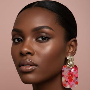 May include: A statement earring featuring a rectangular pendant with a multi-colored confetti pattern in shades of pink, red, and white. The pendant is attached to a textured, gold-toned metal top piece with a hammered finish and a post backing.