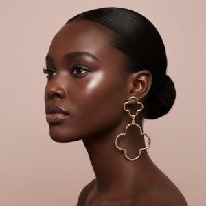 May include: A pair of gold-toned drop earrings featuring two connected open-work clover shapes. The smaller clover sits at the earlobe, while the larger clover hangs below, both constructed from thin, polished metal wire in a four-leaf design.