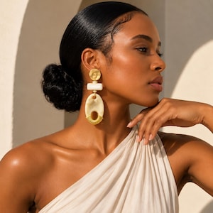 May include: A statement earring featuring a textured gold circular top, a rectangular white bead, and a large oval cream-colored pendant with a ribbed gold metal base. The earring is composed of stacked geometric shapes in gold, white, and cream.