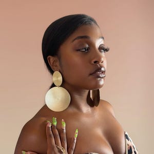 May include: Gold-toned, textured disc earrings. The earrings feature a smaller, circular top piece connected to a larger, round bottom. The image also shows a person wearing the earrings, with long, painted nails and a leopard print top.