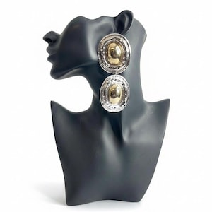 May include: A pair of drop earrings featuring two stacked oval components. Each component has a polished gold-tone center surrounded by a textured, silver-tone border with a hammered finish. The earrings are displayed on a black mannequin bust.