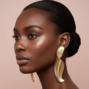 May include: A pair of large, gold-toned earrings featuring a smooth, elongated, and curved abstract shape with a textured, matte finish. The earrings hang vertically from the earlobe, displaying a fluid, ribbon-like design that catches the light.