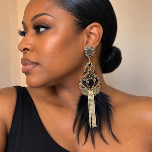 May include: A long, ornate chandelier earring featuring a gold-tone metal frame encrusted with small black crystals. The design includes a central teardrop-shaped black stone, dangling gold-tone crystal fringe, and soft black feathers at the base.
