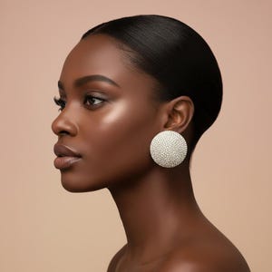 May include: A profile view of a person wearing large, round, silver stud earrings. The earrings are covered in small, sparkling crystals. The person has dark skin and black hair pulled back. The background is a solid, neutral color.