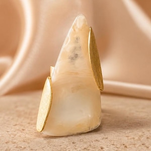 May include: A unique ring featuring a large, triangular, cream-colored stone with gold-colored accents. The stone has a marbled appearance and is flanked by two gold-colored, leaf-shaped elements. The ring is set on a neutral-toned surface.