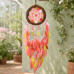 May include: A circular dream catcher featuring a brown twig frame, a central crocheted floral mandala in shades of pink and coral, and hanging strands adorned with pink and lime green beads, finished with long coral and lime green feathers.