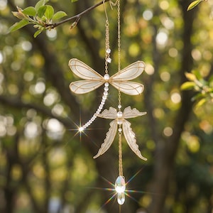 May include: A hanging ornament featuring a gold wire dragonfly with translucent white wings and a beaded tail, connected by a gold chain to a smaller gold butterfly and two clear, faceted crystal teardrop pendants that catch the light.