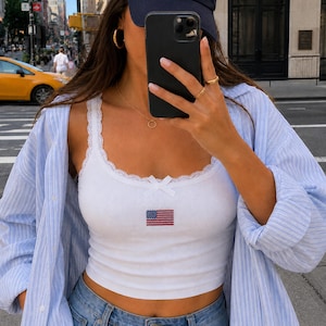 May include: A white ribbed vest top featuring a small embroidered American flag on the chest, a delicate lace trim along the neckline, and a small centre bow. The garment is worn under a light blue and white pinstriped button-down shirt.