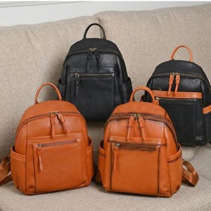 May include: Four leather backpacks in the image. Two are a warm brown color, and two are black. Each backpack has a top handle, a front zippered pocket, and side pockets. The backpacks are sitting on a beige surface.