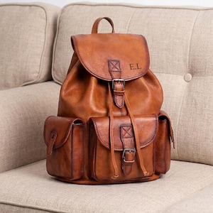 Pode incluir: Uma mochila de couro castanho-claro com uma aba superior com fecho de fivela, dois bolsos laterais e um bolso frontal central com fivela. As iniciais E.L. estão gravadas na aba. A mochila tem um fecho de cordão e uma alça superior.