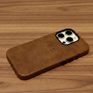 Op de afbeelding: Een bruine lederen telefoonhoes voor een iPhone met een drievoudige camera-opstelling. De achterkant van de hoes is voorzien van de in reliëf aangebrachte initialen E.L. onder de camera-uitsparing. De hoes heeft een gladde textuur en bedekt de achterkant.
