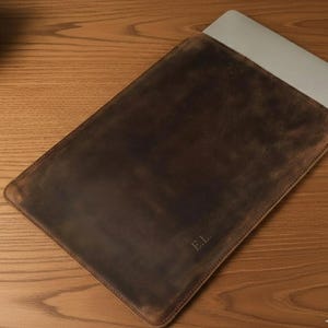 Peut inclure: Une housse pour ordinateur portable en cuir marron foncé avec une finition mate lisse et des coutures visibles sur les bords. Le coin inférieur droit présente les initiales E.L. gravées. Un ordinateur argenté dépasse de l'ouverture.