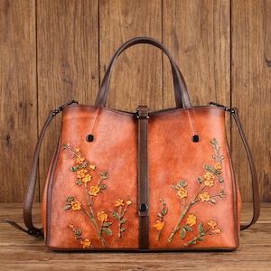 Puede incluir: Un bolso estructurado de cuero color canela con una correa central vertical y cierre a presión. El exterior está decorado con ramas florales amarillas en relieve y pintadas con tallos y hojas verdes, complementado con asas de color marrón oscuro.