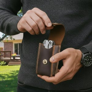 Op de afbeelding: Een donkerbruin lederen etui met zichtbare stiksels en een messingkleurige drukknoopsluiting, met daarin een zilverkleurige multitool. Het etui heeft een klep aan de bovenzijde en een gestructureerd ontwerp voor het opbergen van het gereedschap.