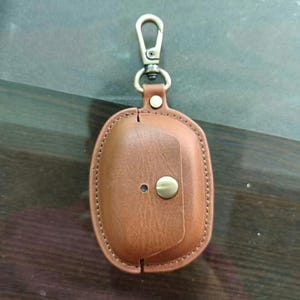 Könnte beinhalten: Eine braune Schutzhülle aus Leder für kabellose Ohrhörer mit einem Klappverschluss, einem messingfarbenen Metalldruckknopf, einer kleinen kreisförmigen Aussparung und einem passenden drehbaren Karabinerhaken aus Metall an einer Lederschlaufe.