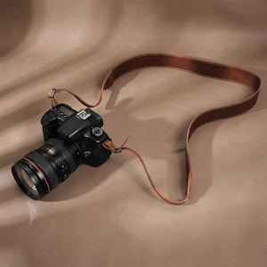 Può includere: Una fotocamera digitale nera con un obiettivo zoom caratterizzato da un sottile anello rosso attorno al barilotto. La fotocamera è fissata a una lunga tracolla in pelle marrone liscia che si curva in un'ampia asola, con fibbie in metallo.