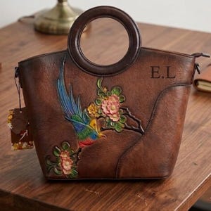 以下が含まれることがあります： 円形のハンドルが付いた茶色のハンドバッグ。カラフルな鳥と花のエンボス加工とペイントが施されています。バッグは質感のある表面で、ファスナー開閉式です。右上には黒字で「E.L.」の文字がプリントされています。