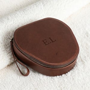 Op de afbeelding: Een bruine leren reisetui met ritssluiting en een klein handvat. De initialen "E.L" zijn op de bovenkant in reliëf aangebracht. De ovale etui ligt op een wit, getextureerd oppervlak.
