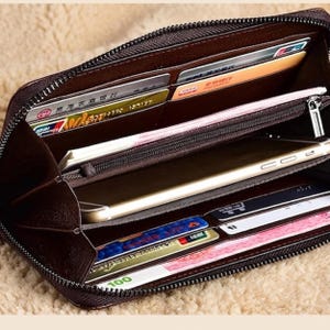 May include: A dark brown zippered wallet featuring multiple interior slots for credit cards, a central zippered compartment, and a dedicated space for a smartphone. The wallet is filled with various cards, paper currency, and a gold-toned mobile device.