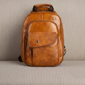 May include: A tan leather backpack featuring a top handle, a main zippered compartment, and a front pocket with a snap button closure. The upper portion of the bag is embossed with the letters E.L. The design includes dark brown straps and zippers.