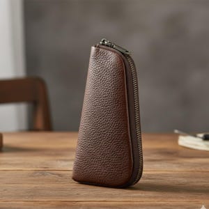 May include: A dark brown, pebble-grained leather key case with a tapered, semi-circular shape. The accessory features a metallic zipper closure that runs along the curved edge, providing a secure opening for the contents stored inside the pouch.