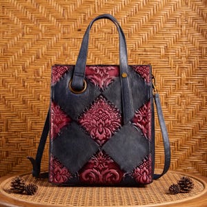 May include: A vertical tote bag featuring a patchwork design of dark navy blue and deep burgundy leather panels. The burgundy sections are embossed with an intricate floral damask pattern. The bag includes two top handles and a shoulder strap.