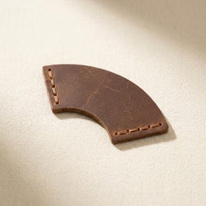 May include: A curved, dark brown leather piece with a smooth, distressed texture. The object features visible light brown stitching along both of its short, straight edges, creating a secure border on this arc-shaped leather component.