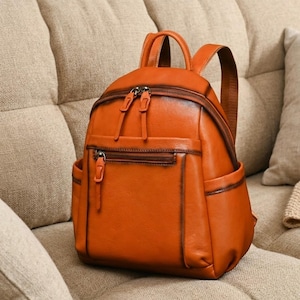 May include: A burnt orange backpack with a smooth finish and dark brown edges. It features a main zippered compartment, a smaller front zippered pocket with a vertical pull tab, two side pockets, a top handle, and adjustable shoulder straps.