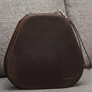 Op de afbeelding: Een donkerbruine, halfstijve, afgeronde driehoekige etui met een gladde matte afwerking. De etui is voorzien van een ritssluiting aan de bovenkant, een bijpassende leren polsband bevestigd aan een zilverkleurige metalen clip en de initialen J.A.
