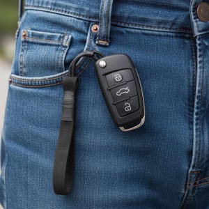 May include: A black car key fob with three buttons featuring icons for locking, trunk release, and unlocking, attached to a black wrist strap. The key fob is hanging from the pocket of a pair of blue denim jeans with visible copper rivets.