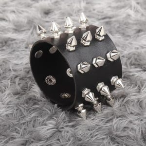 May include: A wide black cuff bracelet featuring three horizontal rows of silver-toned metal spikes. The bracelet is secured with two circular metal snap fasteners visible on the interior side of the band, providing a functional closure mechanism.