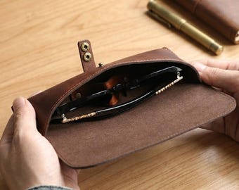 Könnte beinhalten: Ein braunes Brillenetui aus Leder mit einer Umschlagklappe, die durch zwei messingfarbene Metalldruckknöpfe gesichert ist. Das Innere des Etuis ist sichtbar und enthält eine Brille mit schwarzem Rahmen und goldfarbenen Akzenten an den Bügeln.