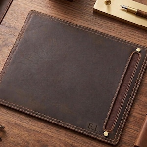 May include: A rectangular dark brown leather desk mat featuring visible stitching along the edges, two brass-colored metal rivets, and the embossed initials E.L. in the bottom right corner. The mat has a smooth surface and a side slot detail.