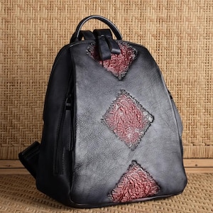 May include: A black backpack featuring a textured surface with three diamond-shaped insets. The insets display a deep red color with an intricate, raised, embossed pattern. The bag includes a top handle, two shoulder straps, and side zippers.