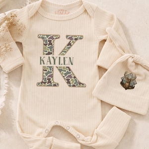 May include: A cream-colored ribbed long-sleeve baby romper with a large letter K featuring a camouflage pattern and the name KAYLEN printed in the center. A matching cream-colored knotted baby hat with a small deer graphic is included.