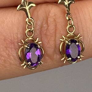 May include: A pair of gold-colored drop earrings. Each earring features a gold floral top, connected to a series of gold loops, and a gold setting holding a purple oval gemstone. The earrings are displayed against a neutral background.