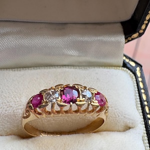 Puede incluir: Un anillo de oro con cinco piedras preciosas. El anillo presenta tres rubíes rojos vibrantes y dos diamantes claros, engastados en una banda calada y decorativa. El anillo se exhibe en un joyero forrado de terciopelo blanco.