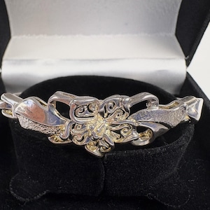 Op de afbeelding: Een zilverkleurige metalen armband met een strik- en bloemmotief, gepresenteerd op een zwart fluwelen oppervlak in een zwarte doos. De armband heeft ingewikkelde details en een gepolijste afwerking, voor sieraden.