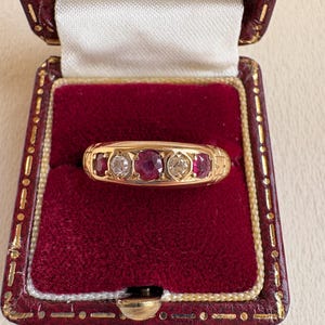 Peut inclure: Une bague en or ornée de trois rubis ovales et de deux petits diamants, sertis dans une monture gravée. La bague est présentée dans un écrin à bijoux vintage en velours rouge avec une fermeture et une bordure dorées.