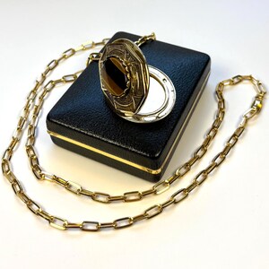 May include: A gold-coloured locket necklace with a rectangular chain. The locket has an octagonal shape with a brown stone in the centre. It rests on a black box with a gold trim.