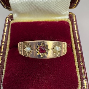 Puede incluir: Un anillo de oro con un rubí central y dos perlas. El anillo presenta un diseño de estrella y se presenta en un joyero de terciopelo rojo.