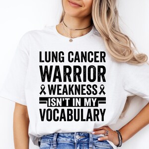 Peut inclure: T-shirt blanc avec l'inscription noire "LUNG CANCER WARRIOR & WEAKNESS ISN'T IN MY VOCABULARY". Le t-shirt est porté avec un jean bleu.