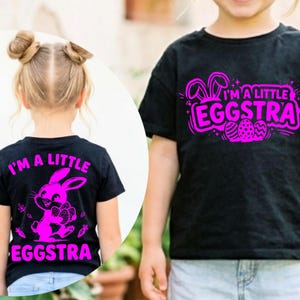 Peut inclure: T-shirt noir avec l'inscription "I'M A LITTLE EGGSTRA" en rose vif. Le motif comprend un lapin, des œufs de Pâques et des oreilles de lapin. Le t-shirt est porté par un jeune enfant.