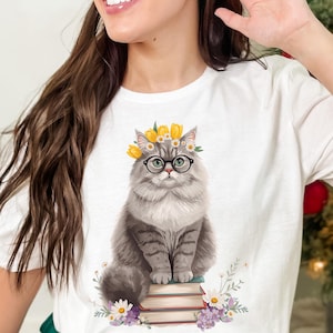 Peut inclure: T-shirt blanc avec une illustration de chat gris portant des lunettes et une couronne de fleurs, assis sur une pile de livres. Le design comprend des fleurs. Un motif charmant pour les amoureux des chats.