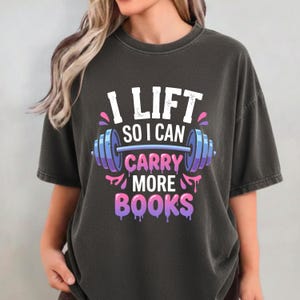 Peut inclure: T-shirt gris foncé avec l'inscription "I LIFT SO I CAN CARRY MORE BOOKS" en blanc, rose et bleu. Le design comprend un haltère. Le t-shirt est de style décontracté et confortable.