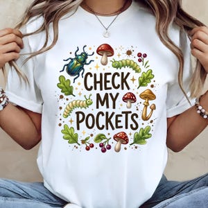 Peut inclure: T-shirt blanc avec un motif graphique coloré représentant un scarabée, des champignons, des chenilles et le texte "CHECK MY POCKETS". Le motif est entouré de feuilles et de baies.